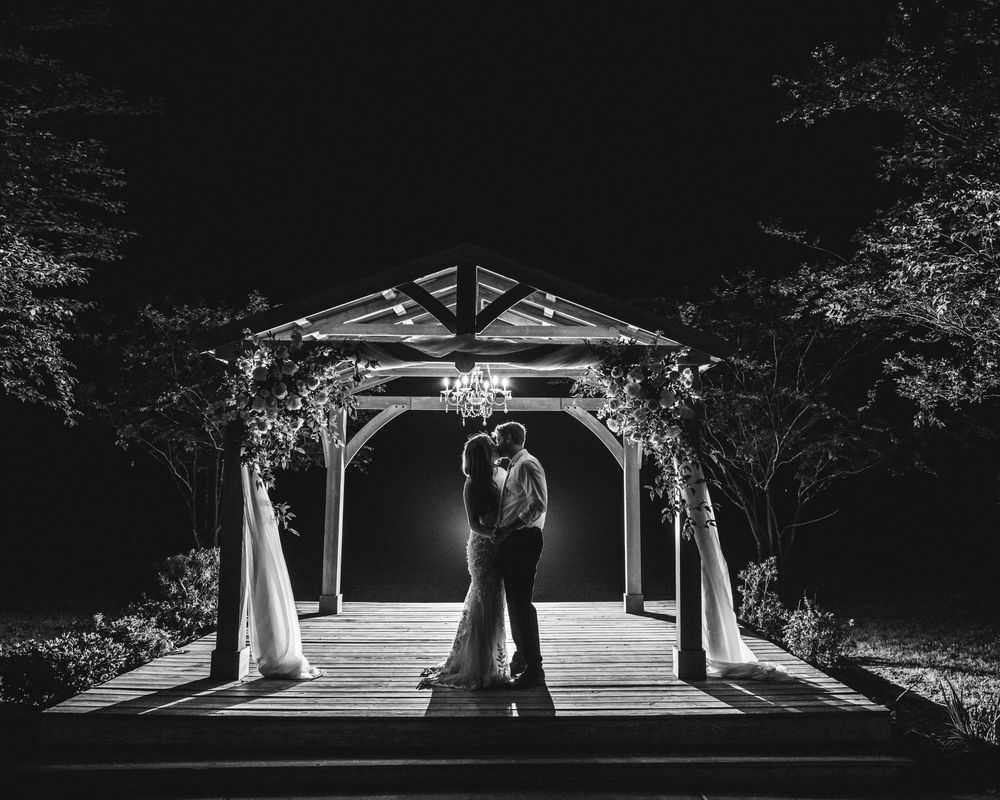 A romantic silhouette of a couple embracing under an illuminated gazebo at night with soft lighting and draped fabric.