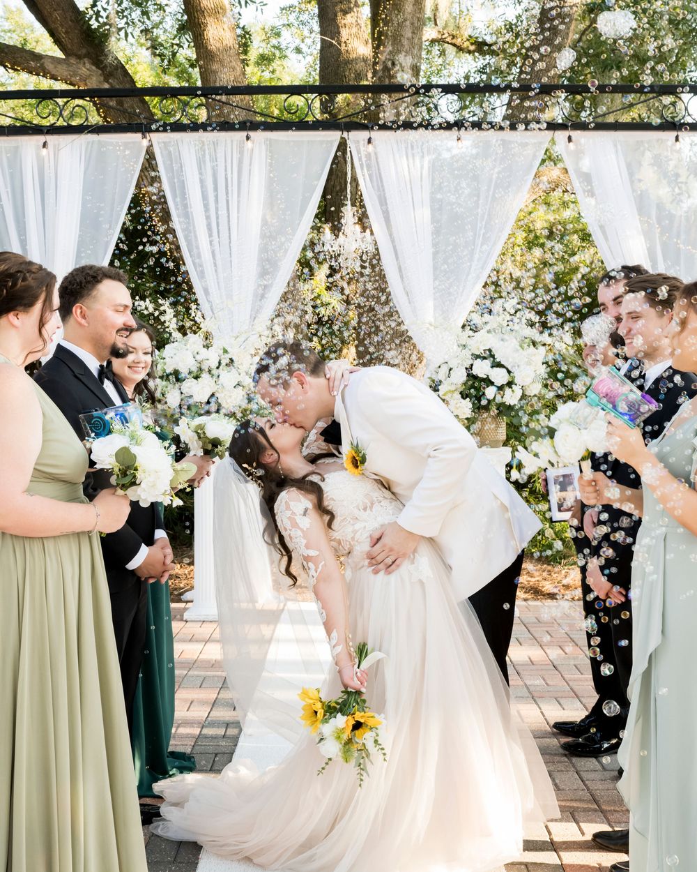 Couple shares intimate first kiss during outdoor wedding ceremony under white draping and natural greenery.