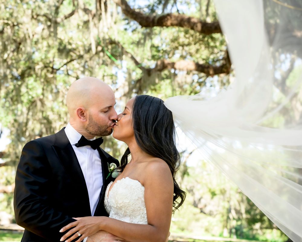 A newlywed couple shares a romantic kiss outdoors under sunlit trees as the bride's veil flows in the breeze.