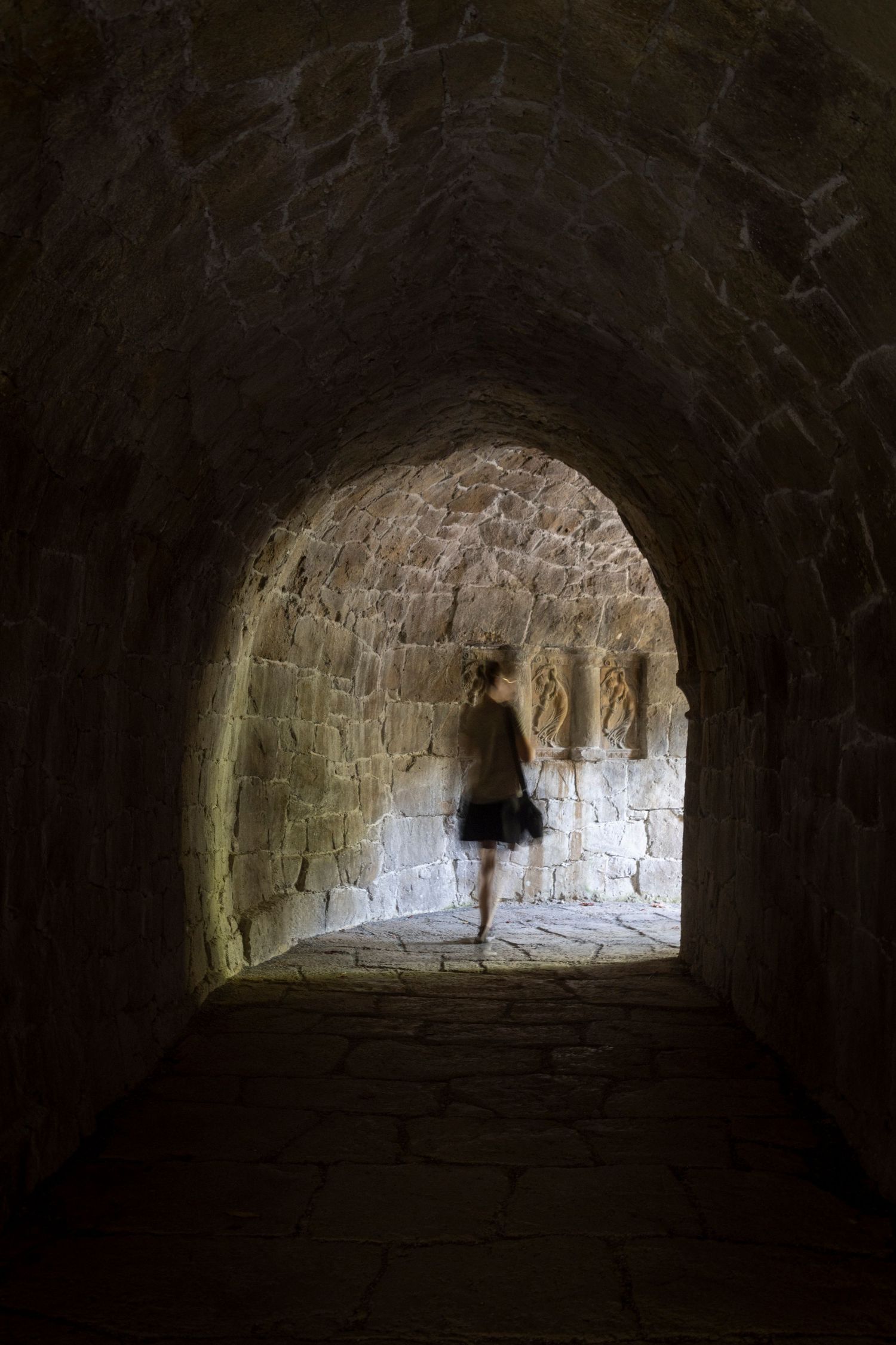 Blurred figure of a woman walking through a stone tunnel into sunlight, creating a moody, dreamlike atmosphere.