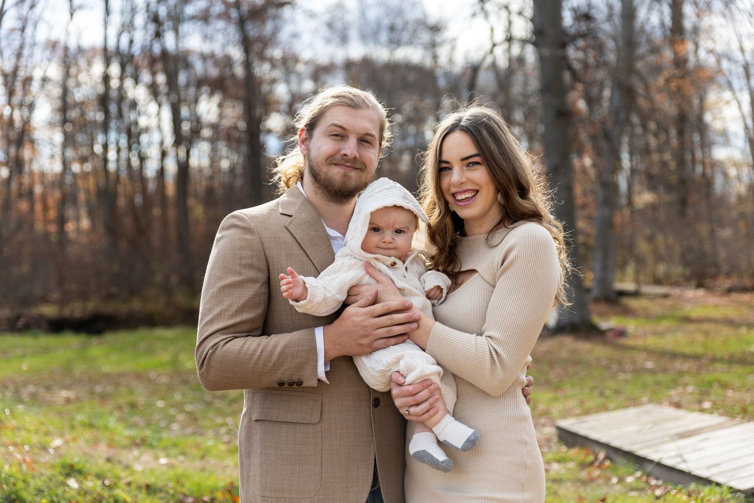 Smiling family of three poses outdoors in autumn, parents holding a baby dressed in white.
