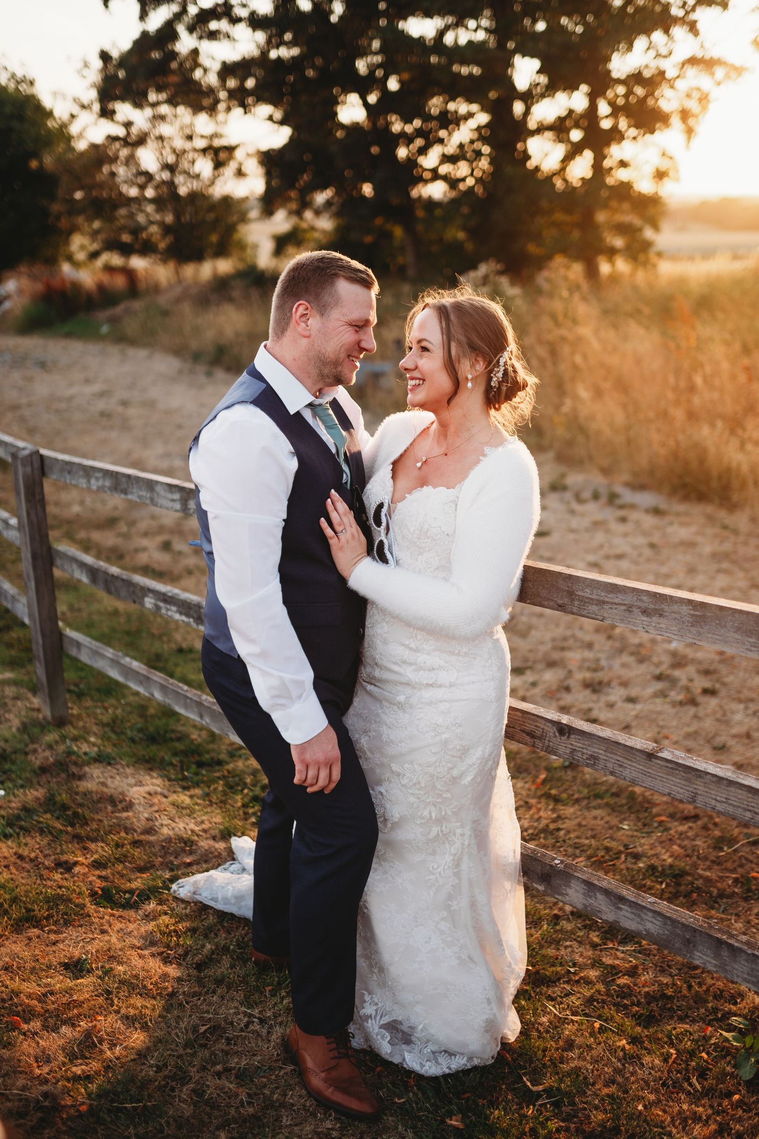 Bride and groom share a romantic moment at golden hour by a rustic fence, bride in lace gown, groom in navy vest.
