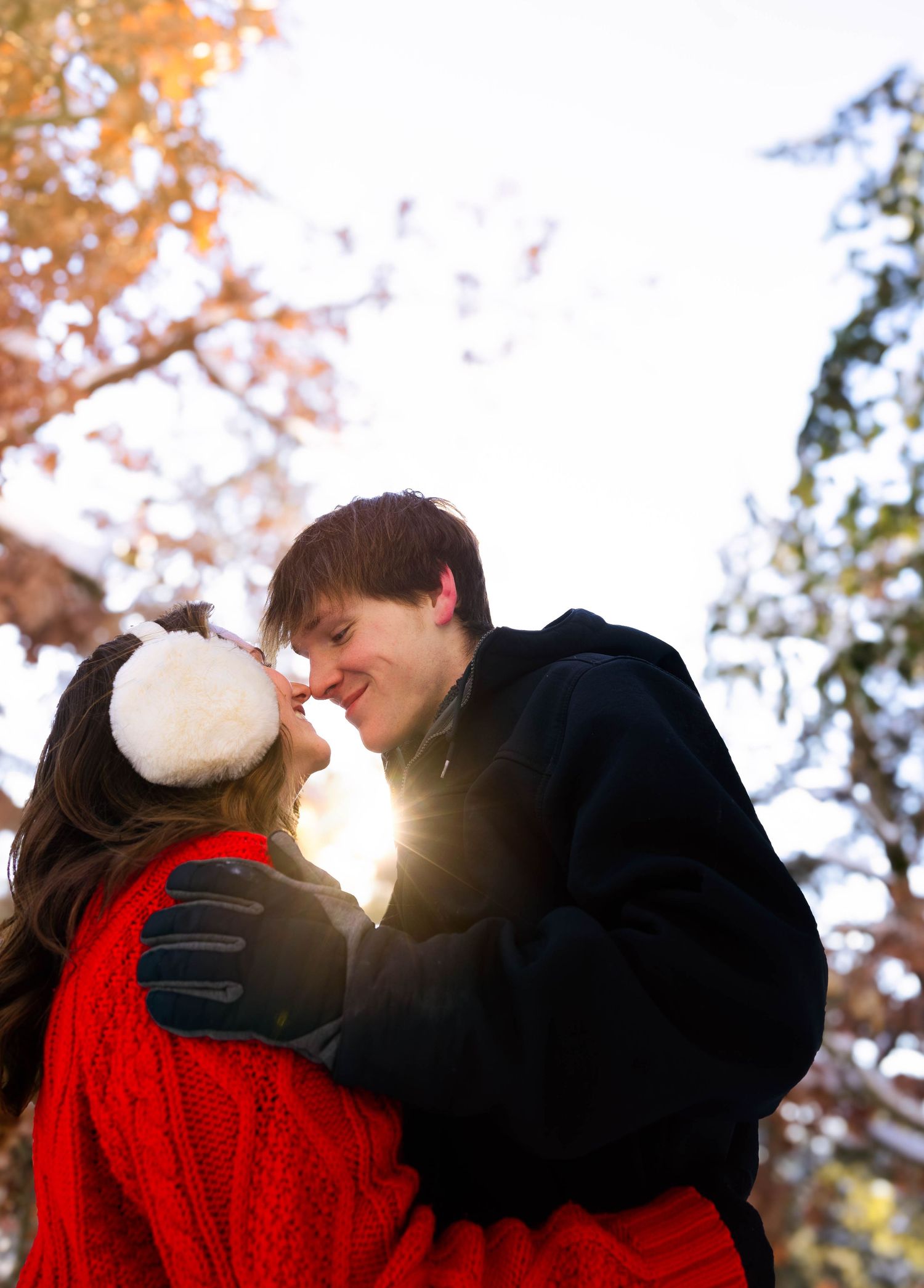 A couple sharing a romantic moment outdoors in winter, with sun flare, snow-dusted trees, and colorful autumn foliage.