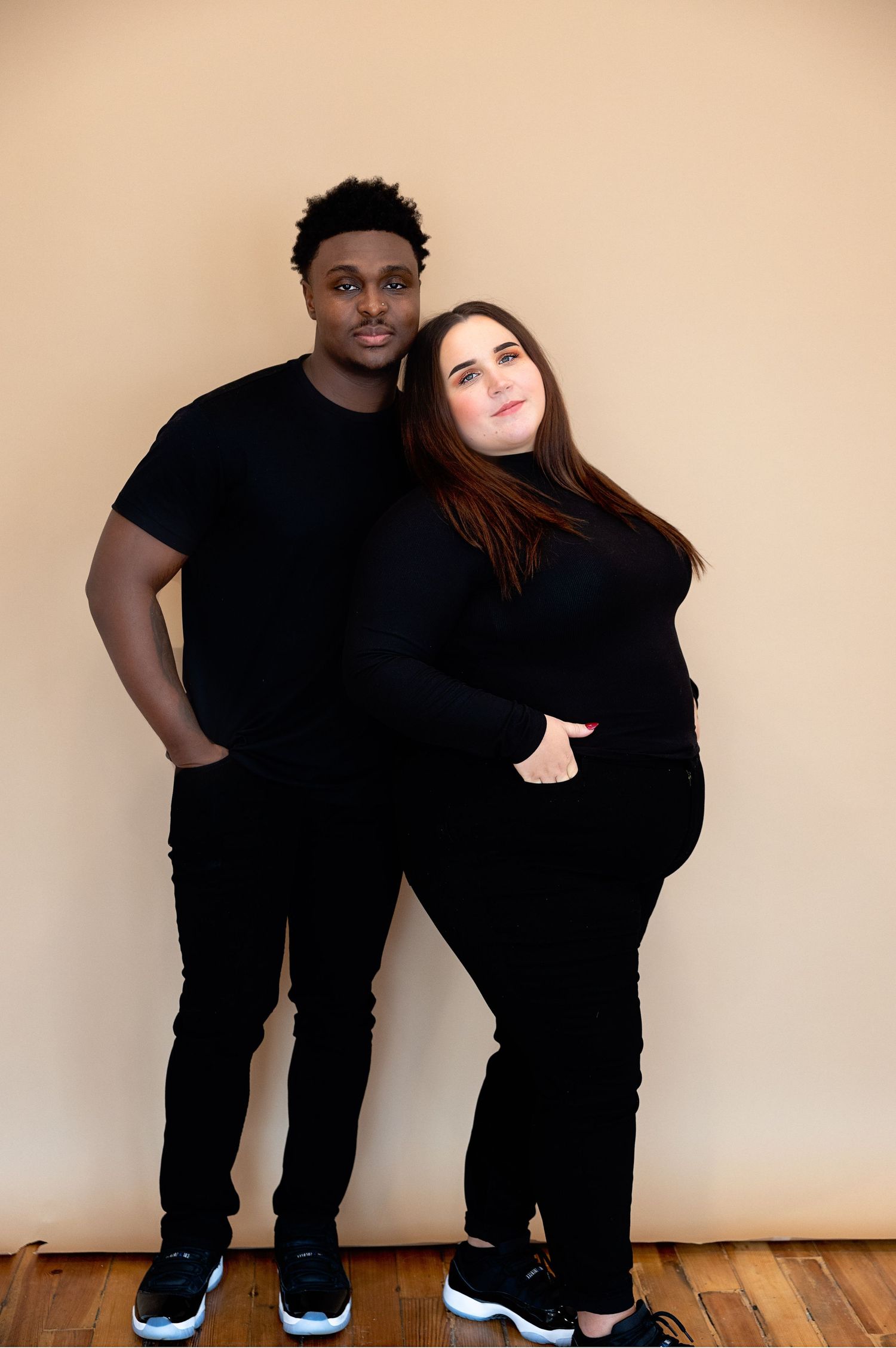 A couple in matching black outfits poses together against a beige backdrop in a studio setting.