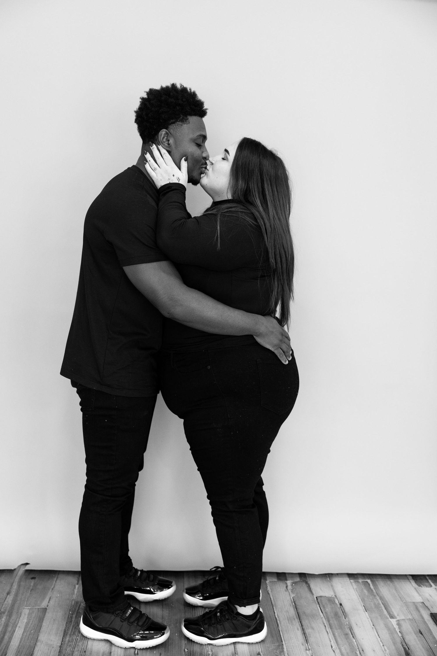 A couple in black outfits shares an intimate moment against a white studio backdrop.