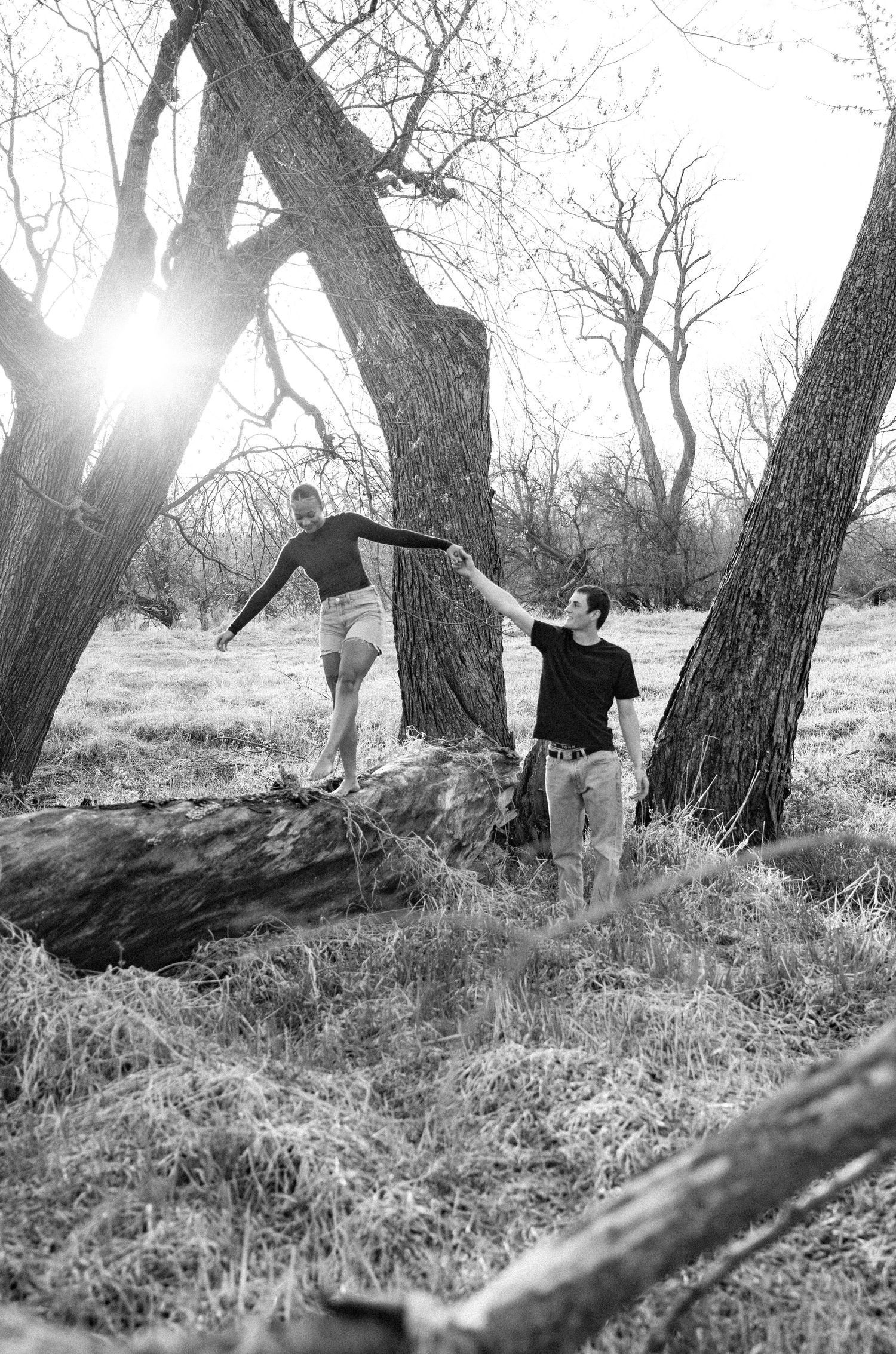 A playful black and white photo of two people having fun near large trees in a field.