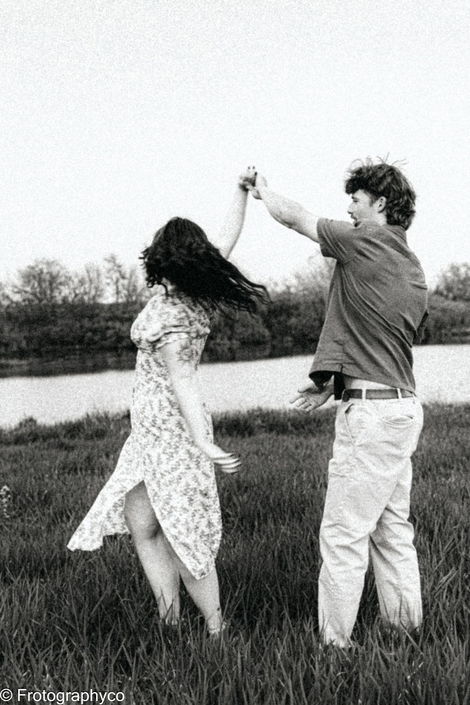 A black and white photo capturing a couple dancing joyfully in a field by the water.