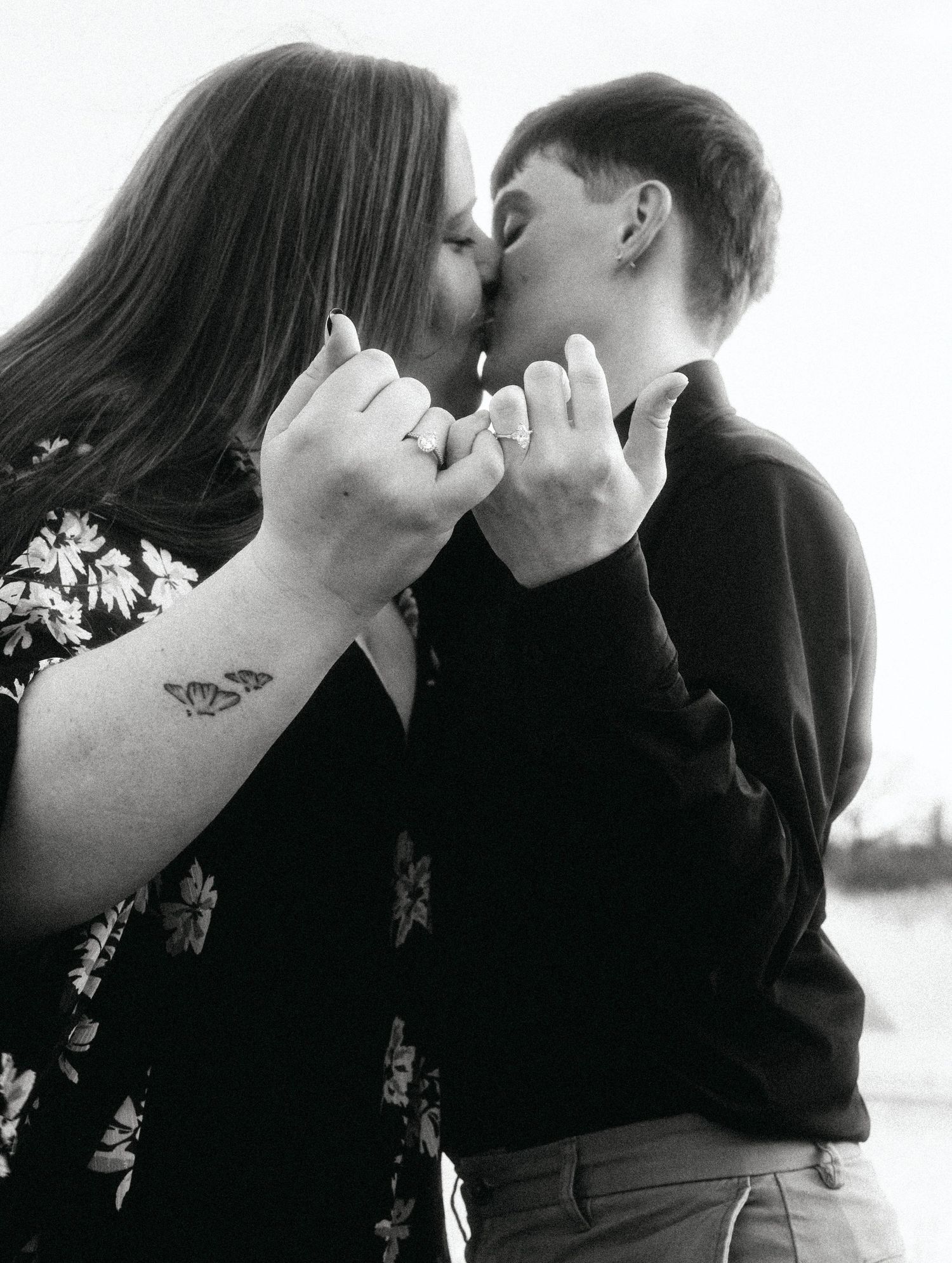 Black and white photo of two people kissing while holding hands, one with a butterfly wrist tattoo.