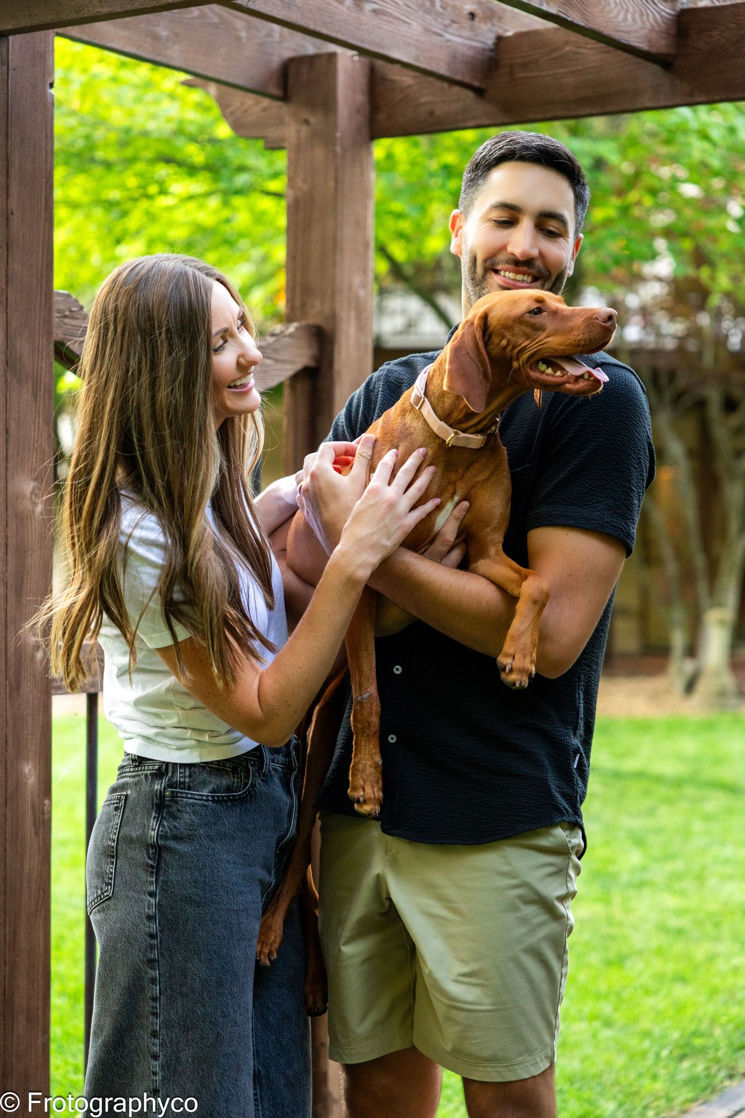 A smiling couple holds their happy Vizsla dog under a wooden pergola in a lush green backyard.