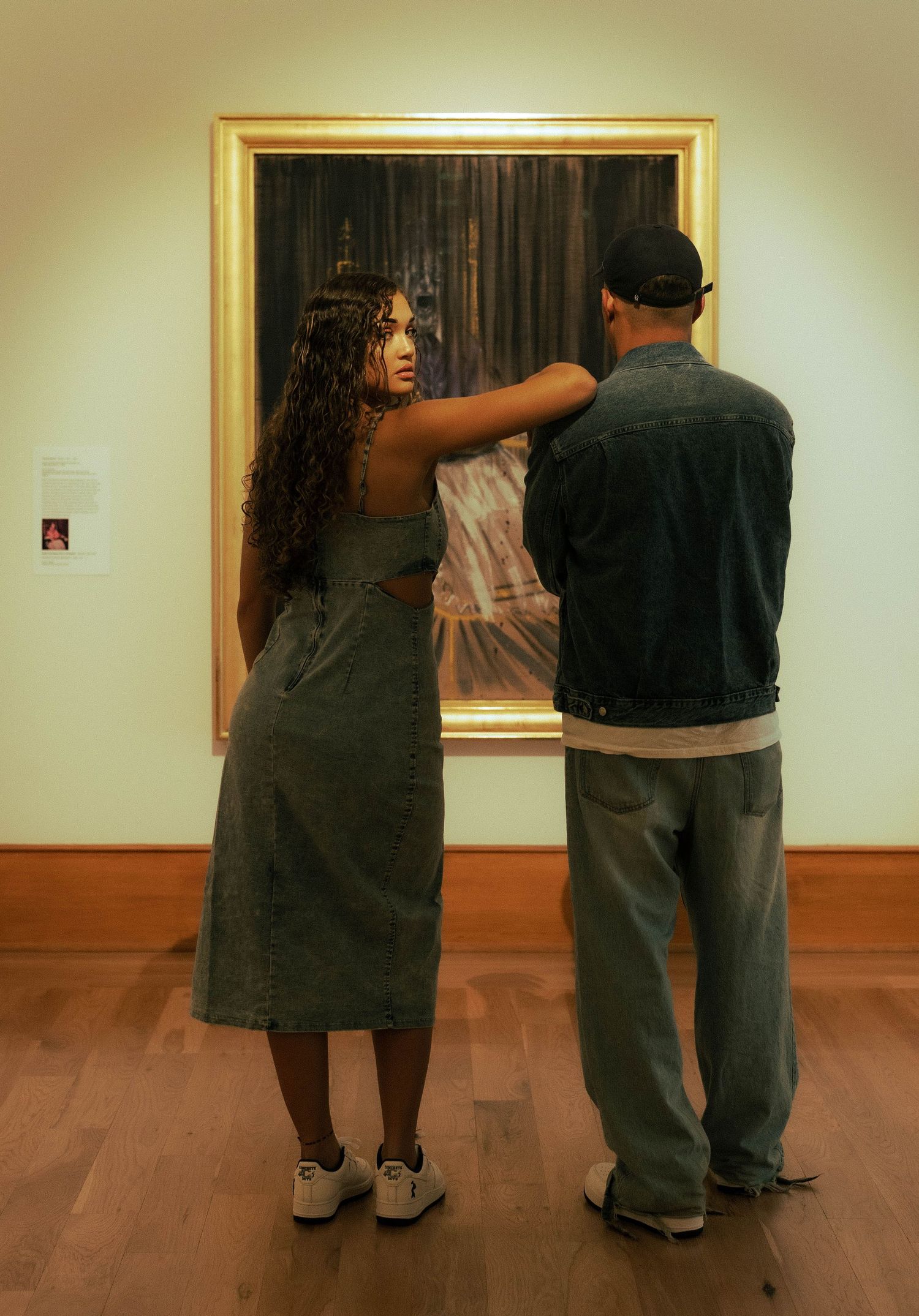 Couple standing together in art museum viewing a large framed painting on a white gallery wall with wood floors.