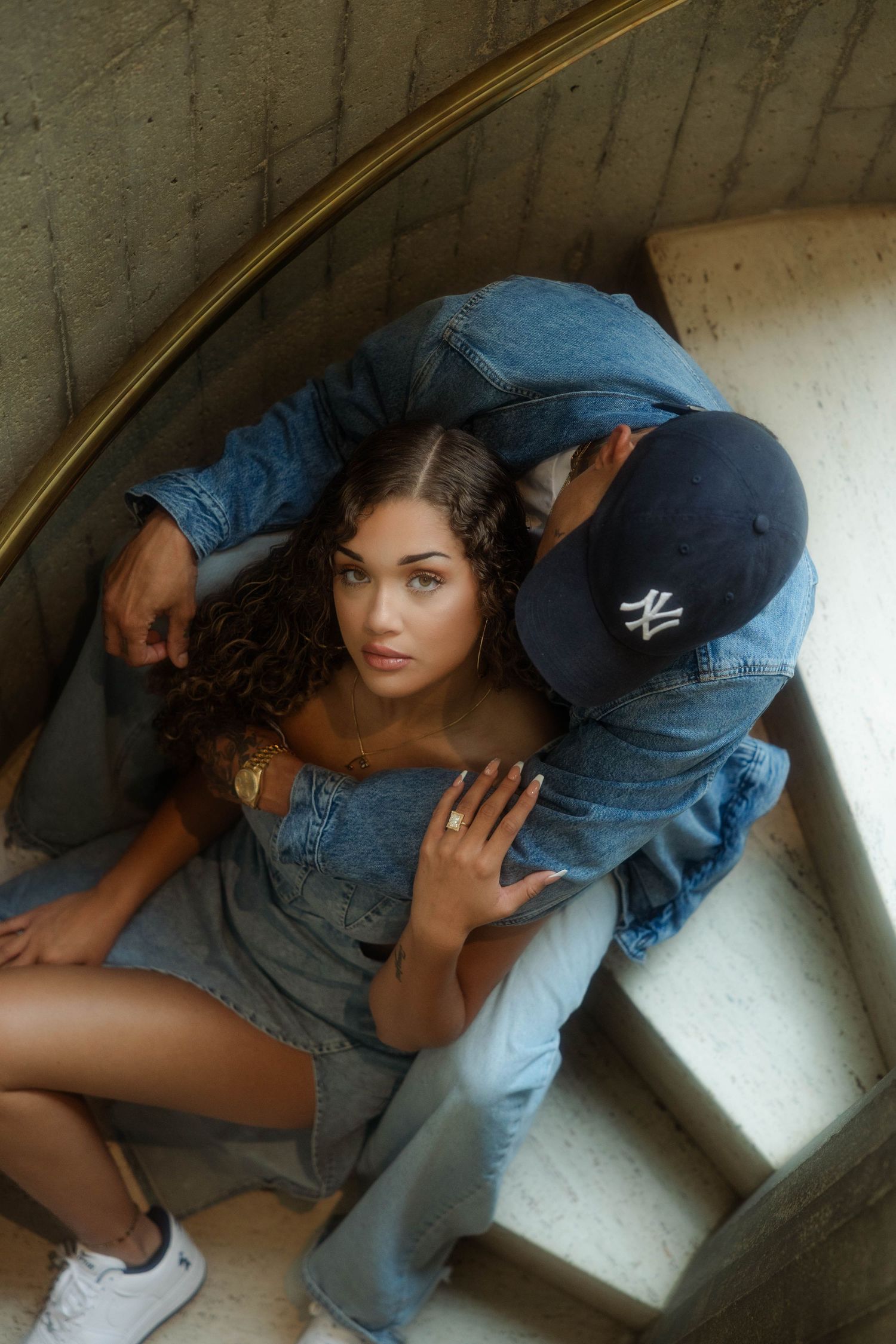 A young woman in denim sits on spiral stairs, leaning against a man in a NY cap and denim jacket.