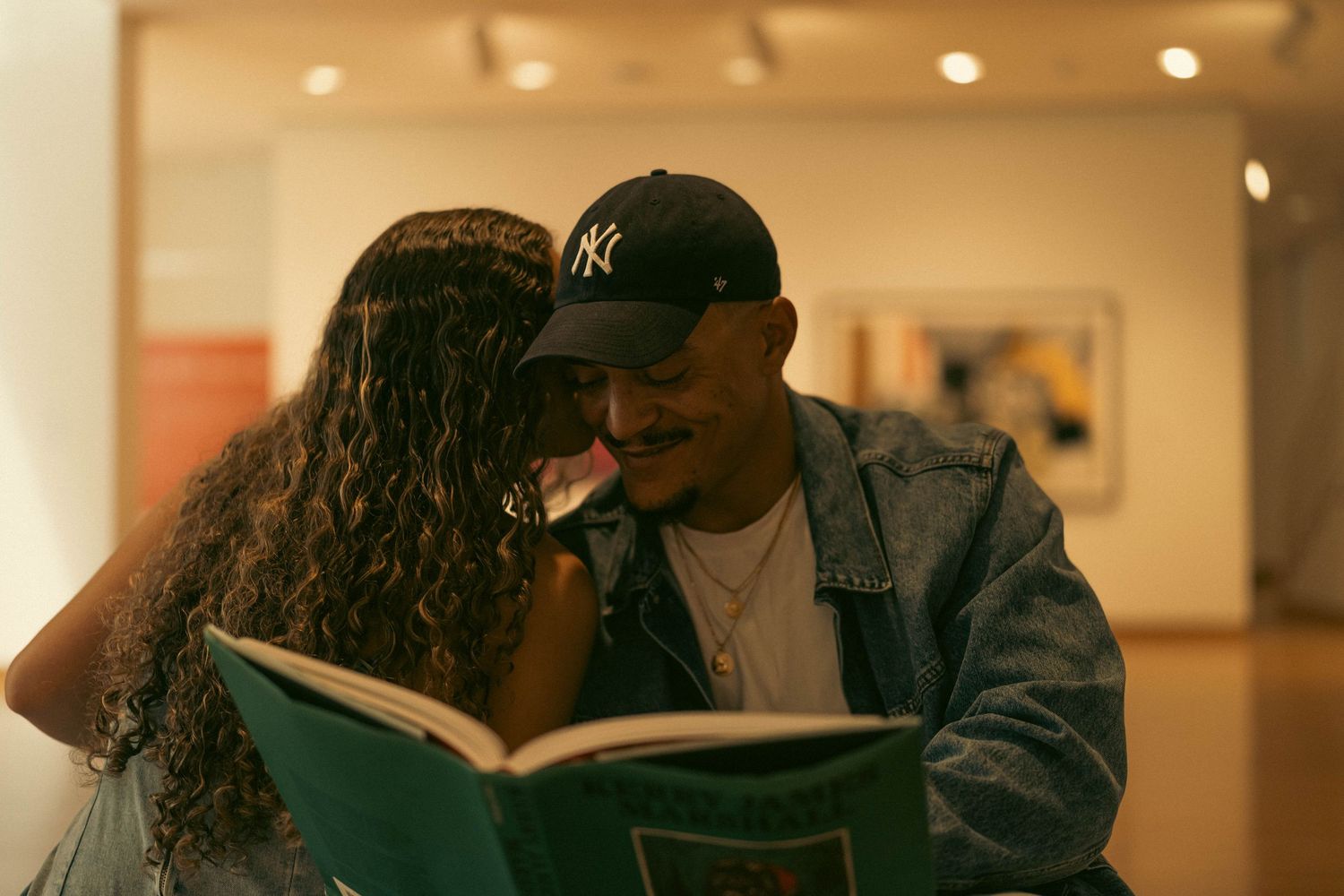 Two people share an intimate moment reading a book together in a warmly lit art gallery.