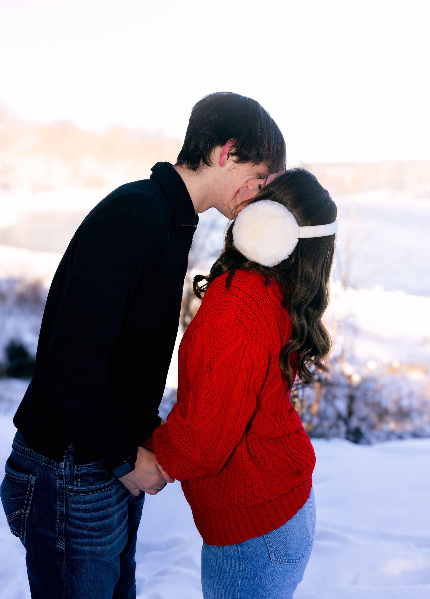 Couple sharing a romantic kiss in the snow, woman wearing red sweater and white earmuffs, winter landscape background.