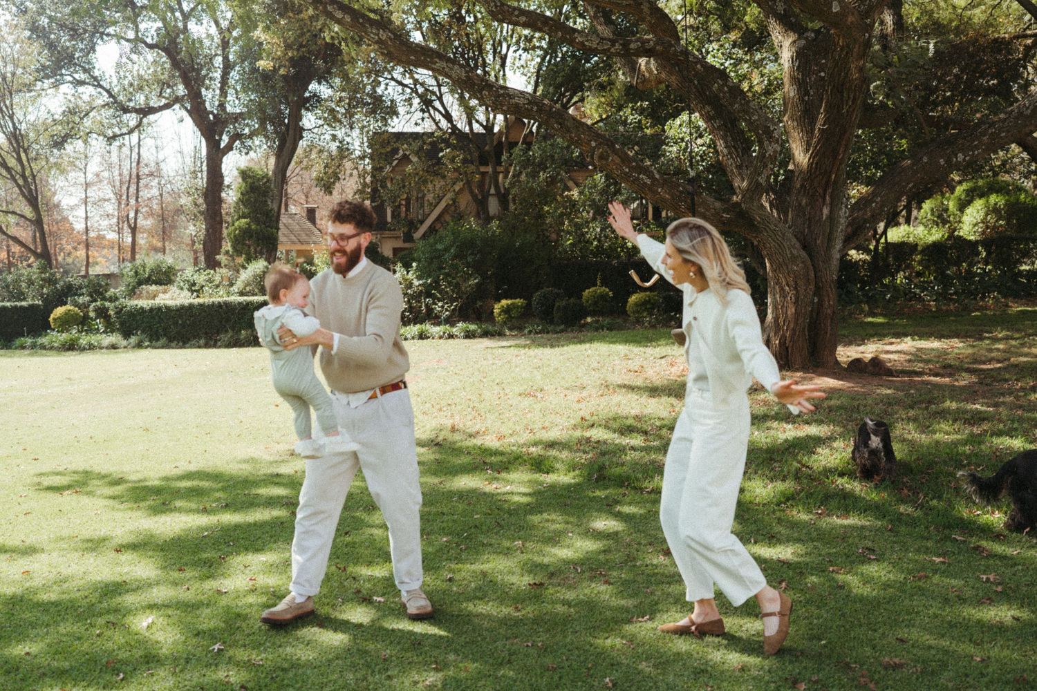 People in white clothing playing and moving across a sunlit grassy area under trees.