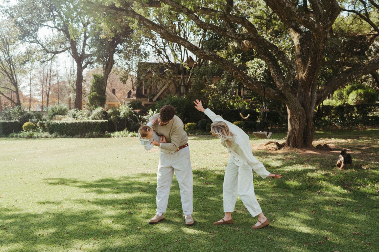 People in light colored outfits enjoying a playful moment on grass under trees.
