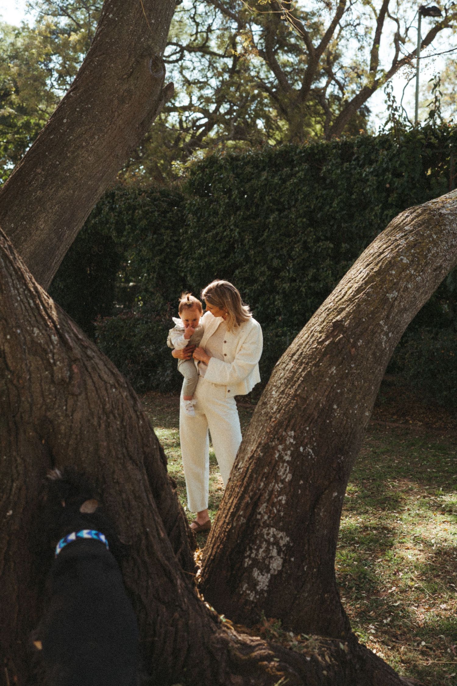 Two people in white outfits share an intimate moment between large tree trunks in a garden.