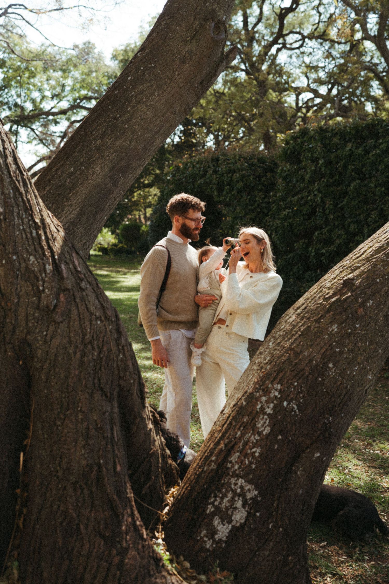 A couple dressed in white shares a playful moment while leaning against a large tree trunk.