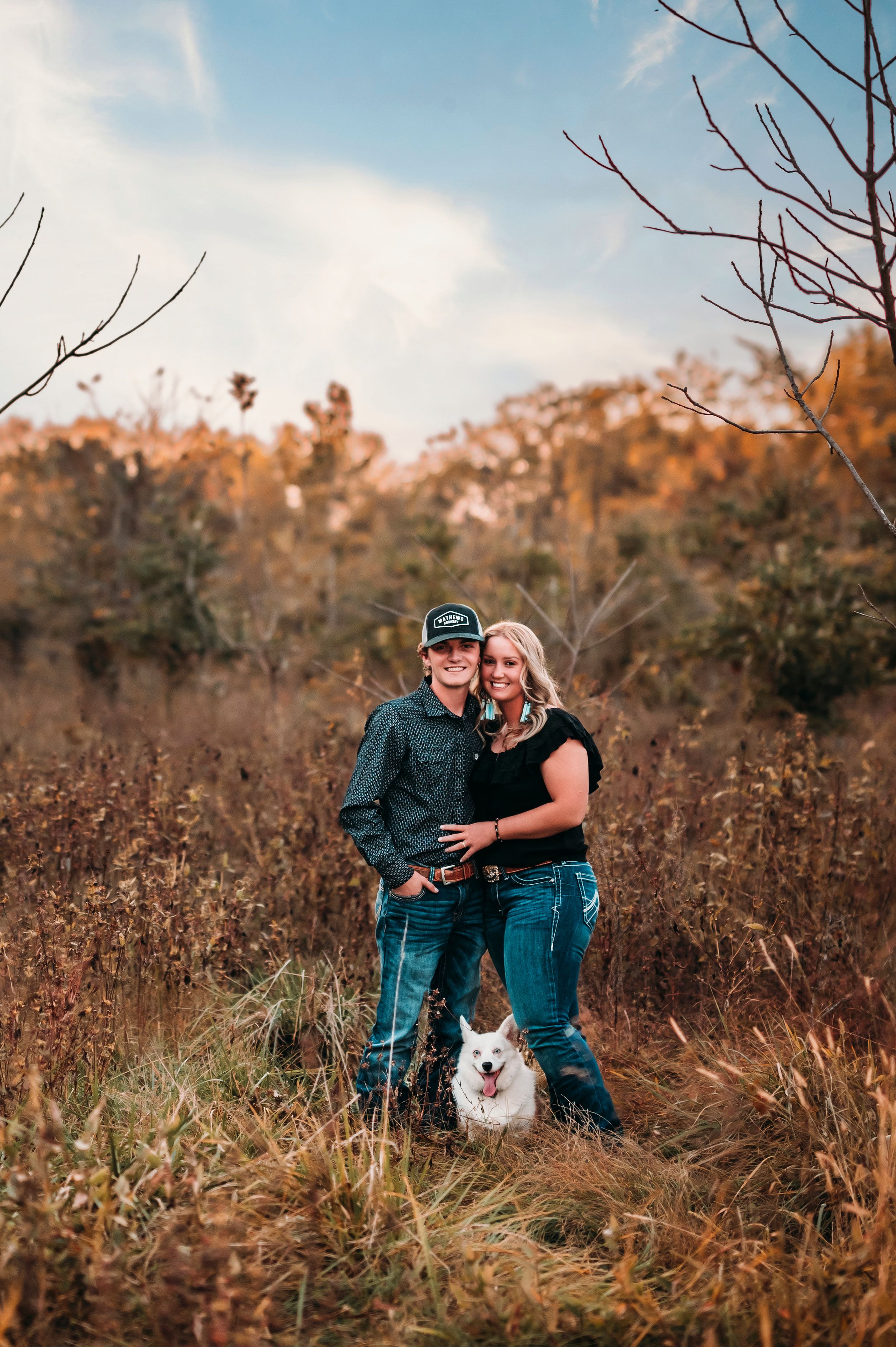 Couple posing with white dog in golden autumn field under blue sky.