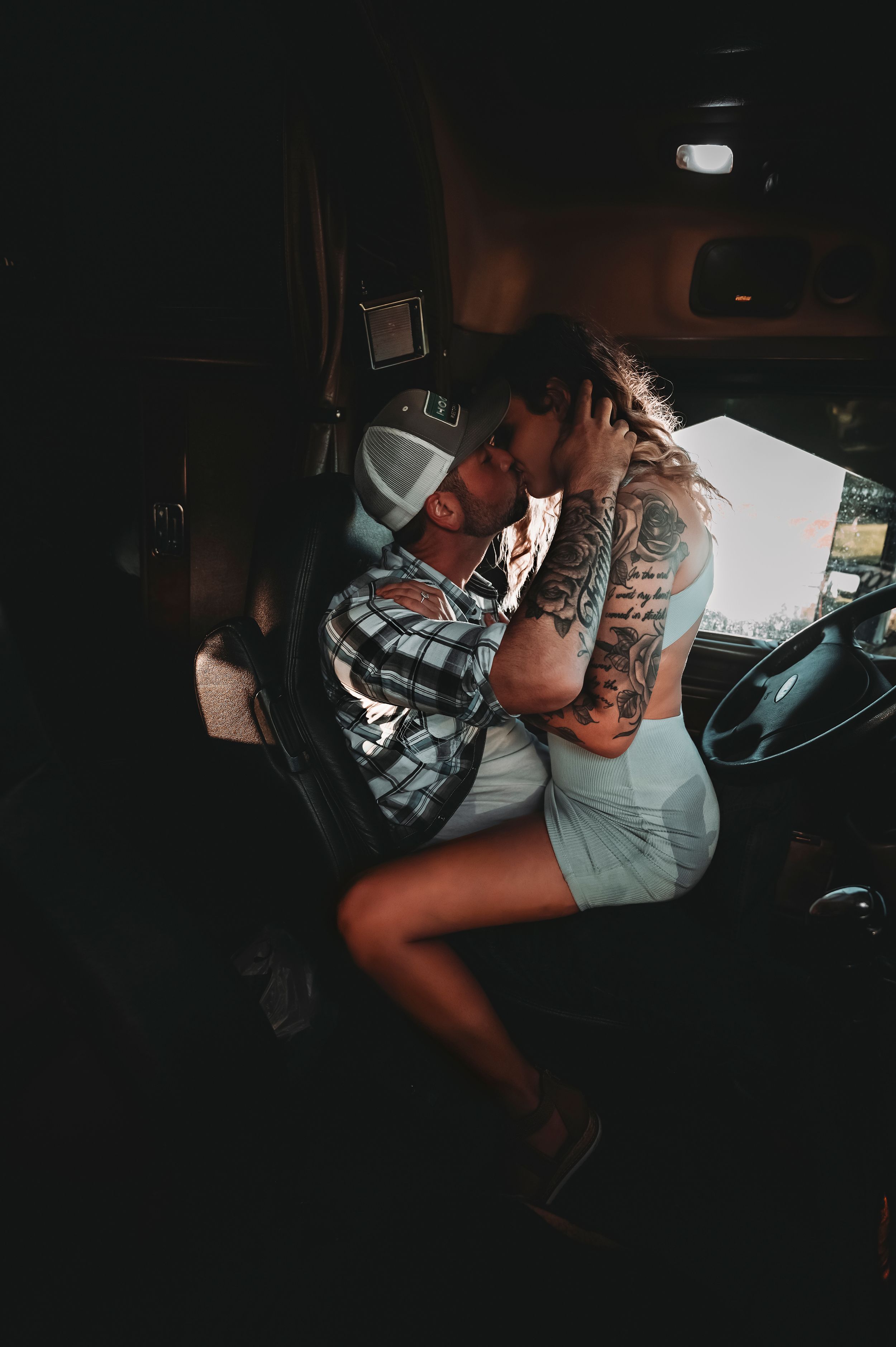 Tattooed couple sharing an intimate kiss inside the cab of a large truck in dim lighting.