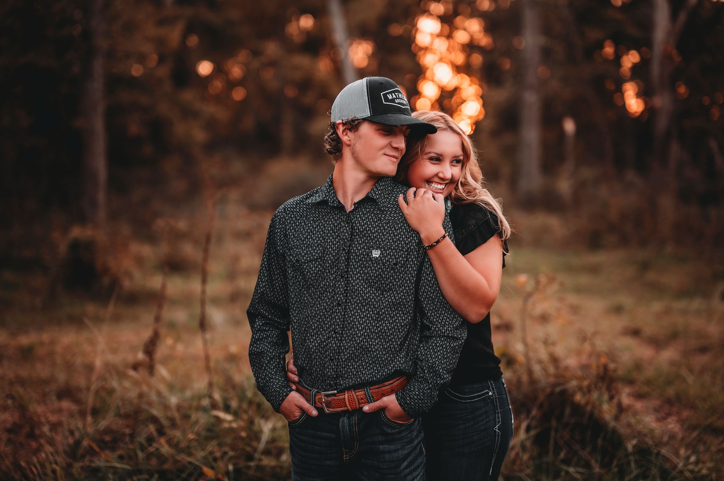 Couple embracing at sunset in a wooded area, man wearing trucker hat.