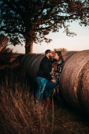 Romantic couple leaning against hay bales in a golden field at dusk beneath a large oak tree.