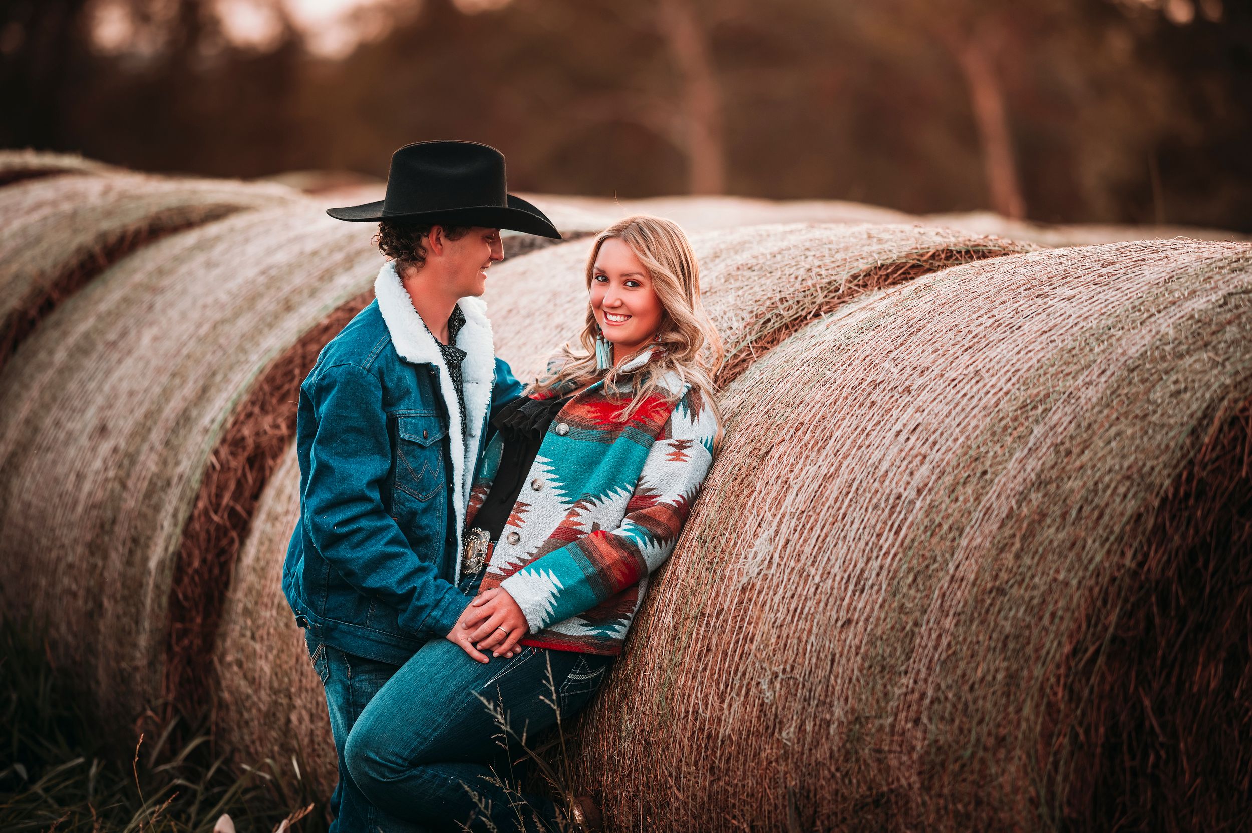 Couple leaning against hay bales at sunset, wearing western attire.