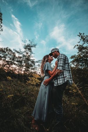 Couple sharing a tender kiss in a lush green meadow beneath a vivid blue sky at sunset.