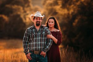 Cowboy couple posing in a warm golden field at sunset, man in plaid shirt and white hat.