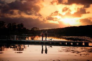 Silhouette of couple and dog on a dock at dramatic sunset with vibrant orange sky reflecting on water.
