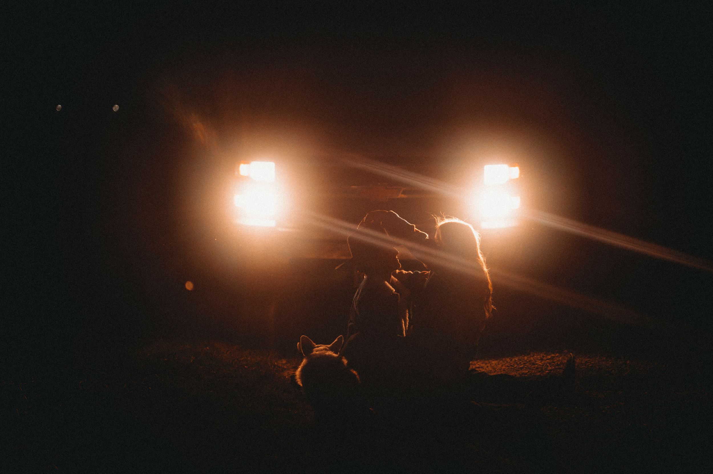 Silhouetted couple sitting between two bright headlights in darkness.