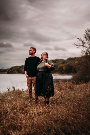 Couple standing in autumn field near lake under dramatic cloudy sky.