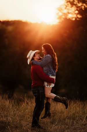 Man lifting woman in cowboy hat during romantic embrace in a glowing golden field at sunset.