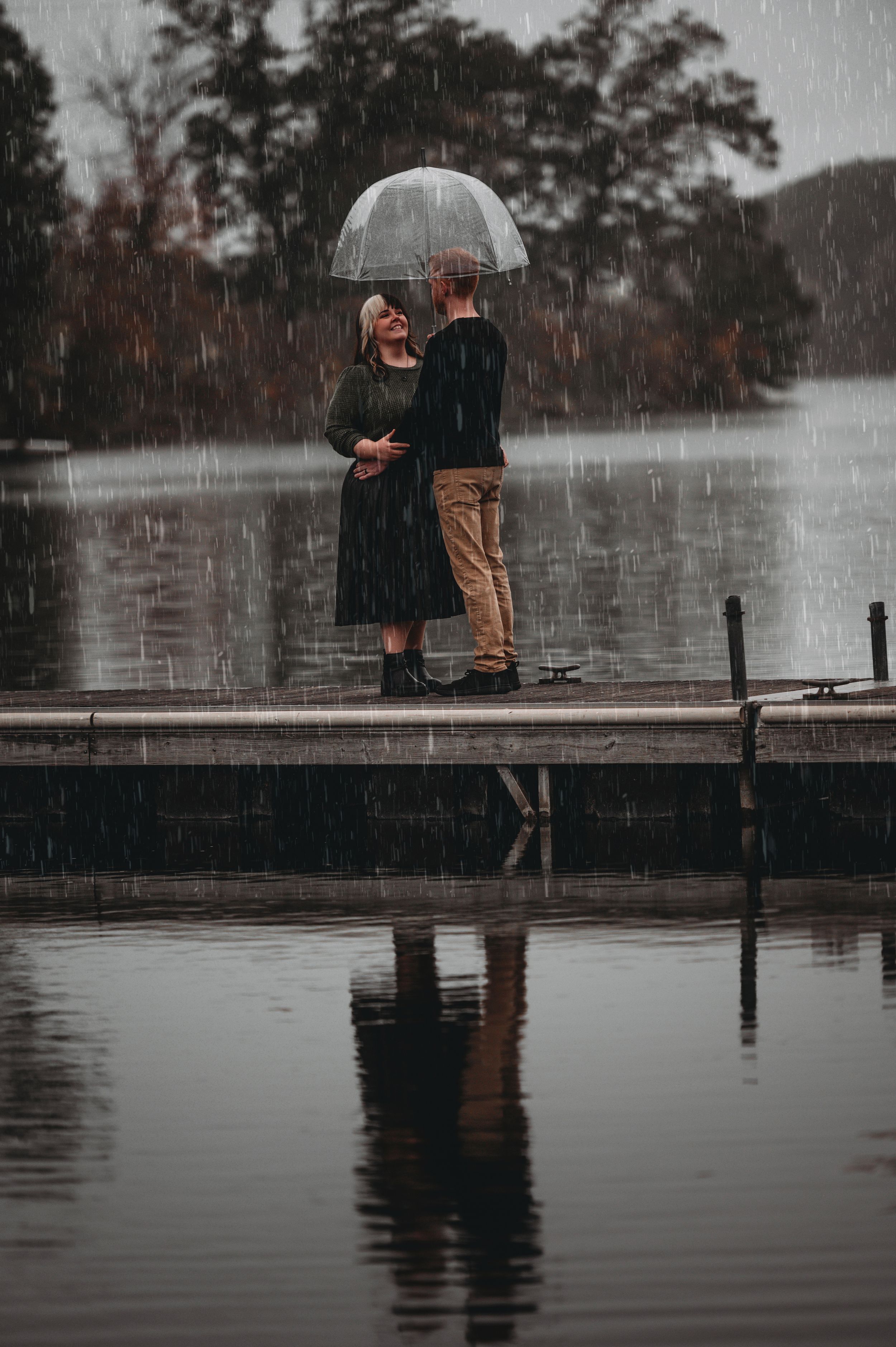 Couple sharing an umbrella on a rainy dock by a lake, their reflection visible in the water below.