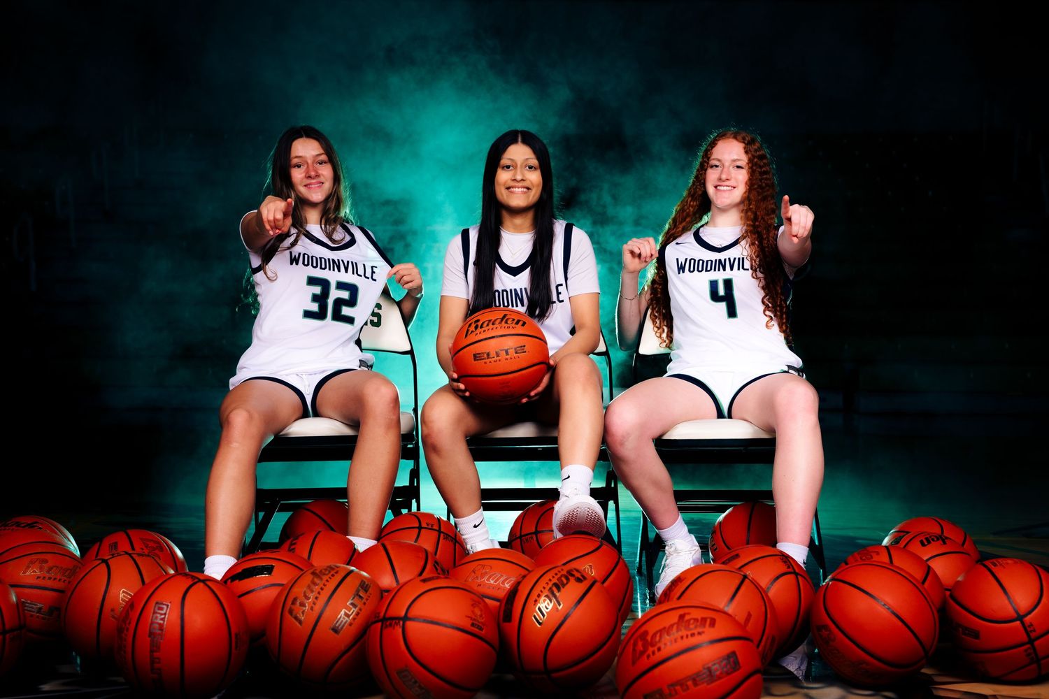 Three basketball teammates pose for a team photo surrounded by basketballs with a turquoise backdrop.
