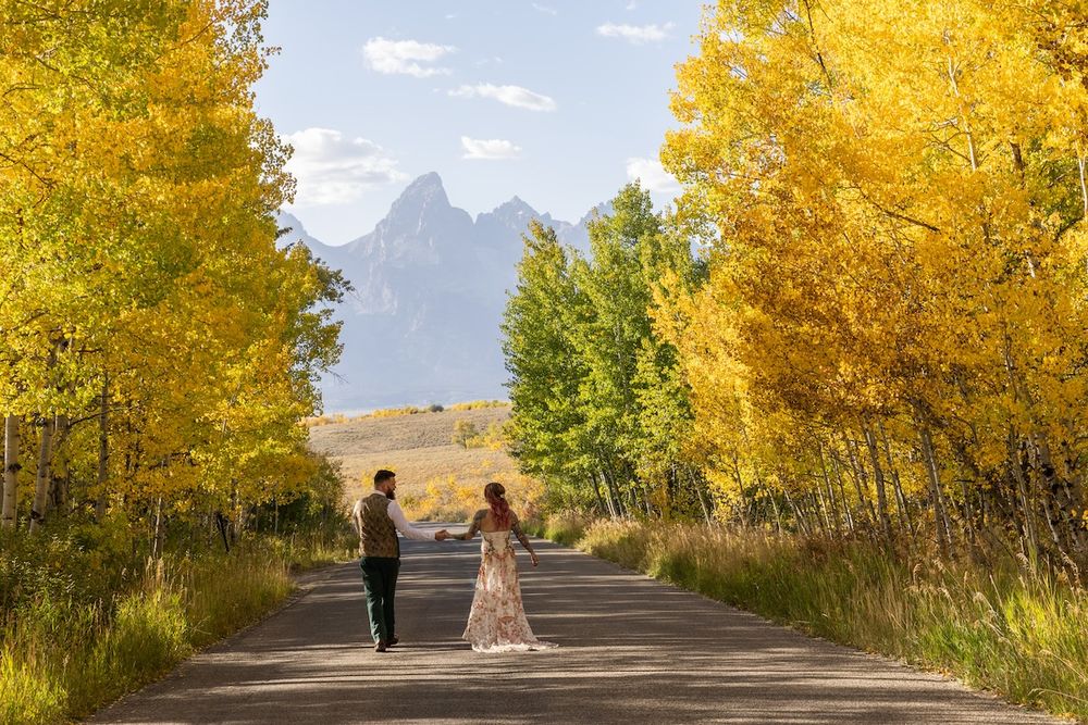 Couple holds hands on a tree-lined road with golden fall foliage and Grand Teton mountains in the background.