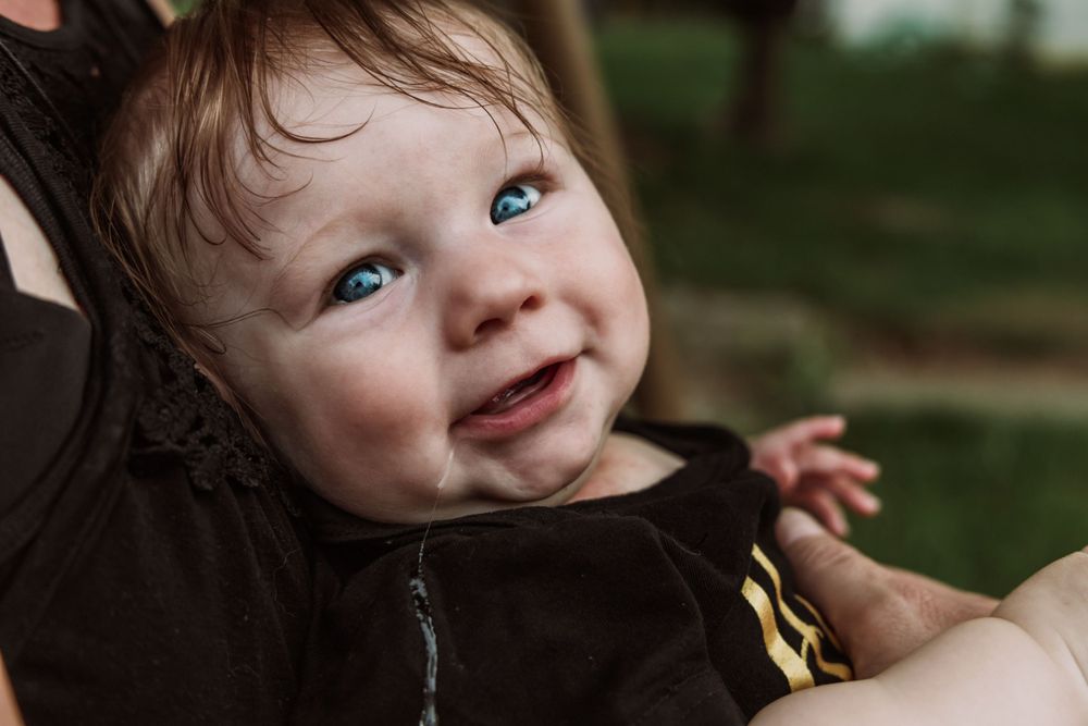 Cheerful baby wearing a brown hoodie smiles with bright blue eyes against a blurred natural background.