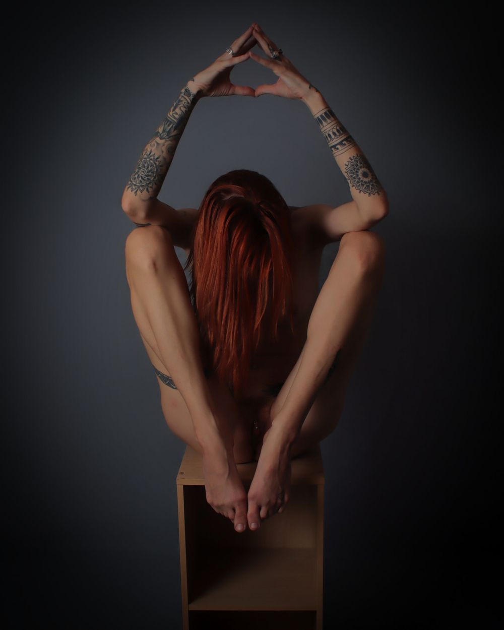 A red haired woman poses on a wooden box in a studio. Her elbows rest on her knees.