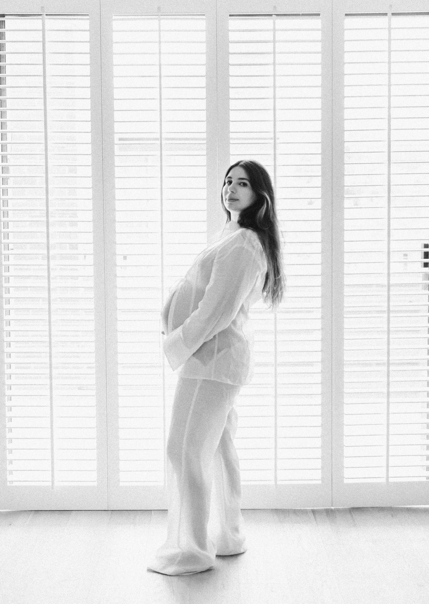 Black and white maternity photo of woman in white outfit standing by window shutters.