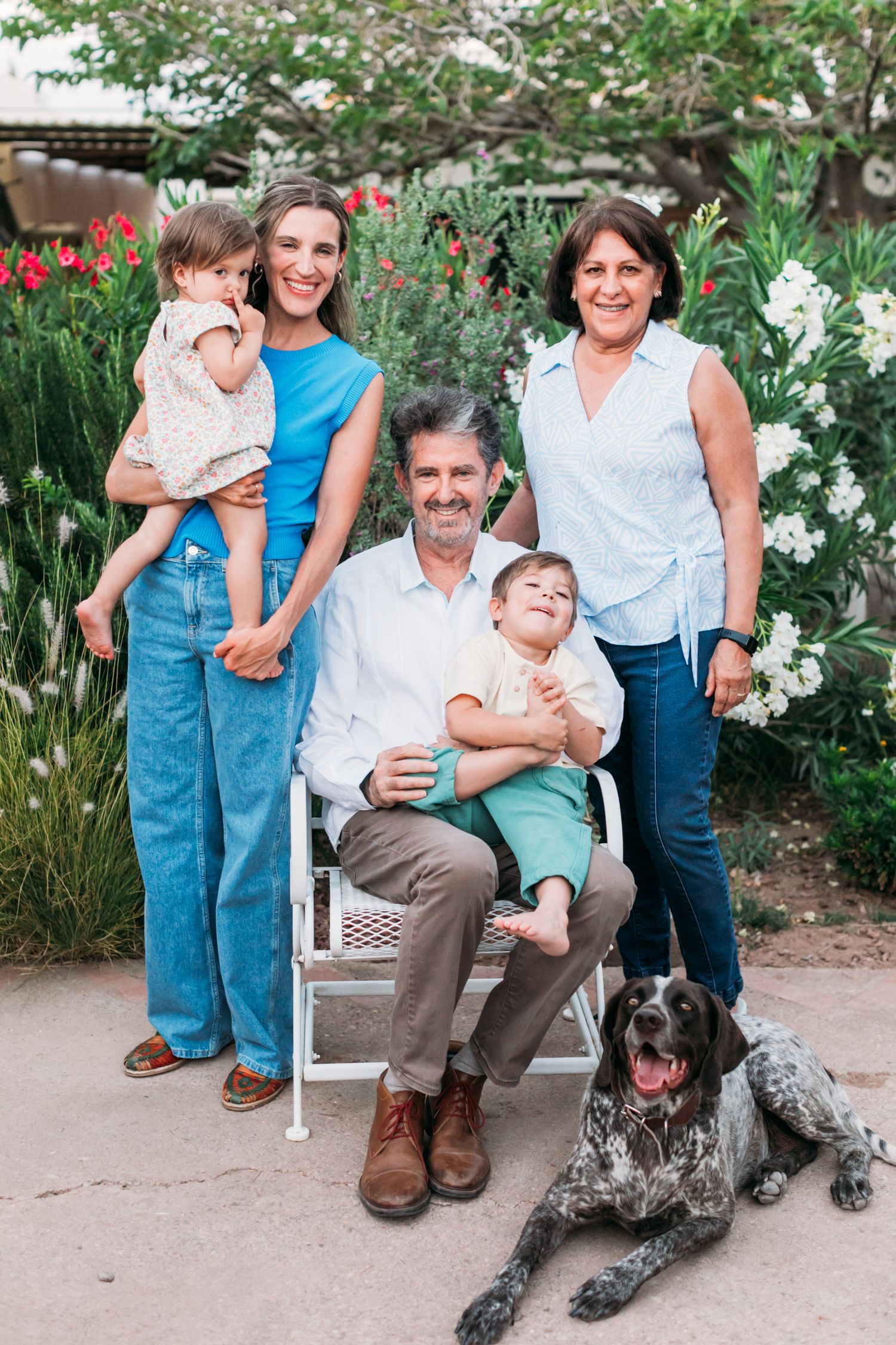 A family portrait in a garden with blooming flowers and a spotted dog resting at their feet.