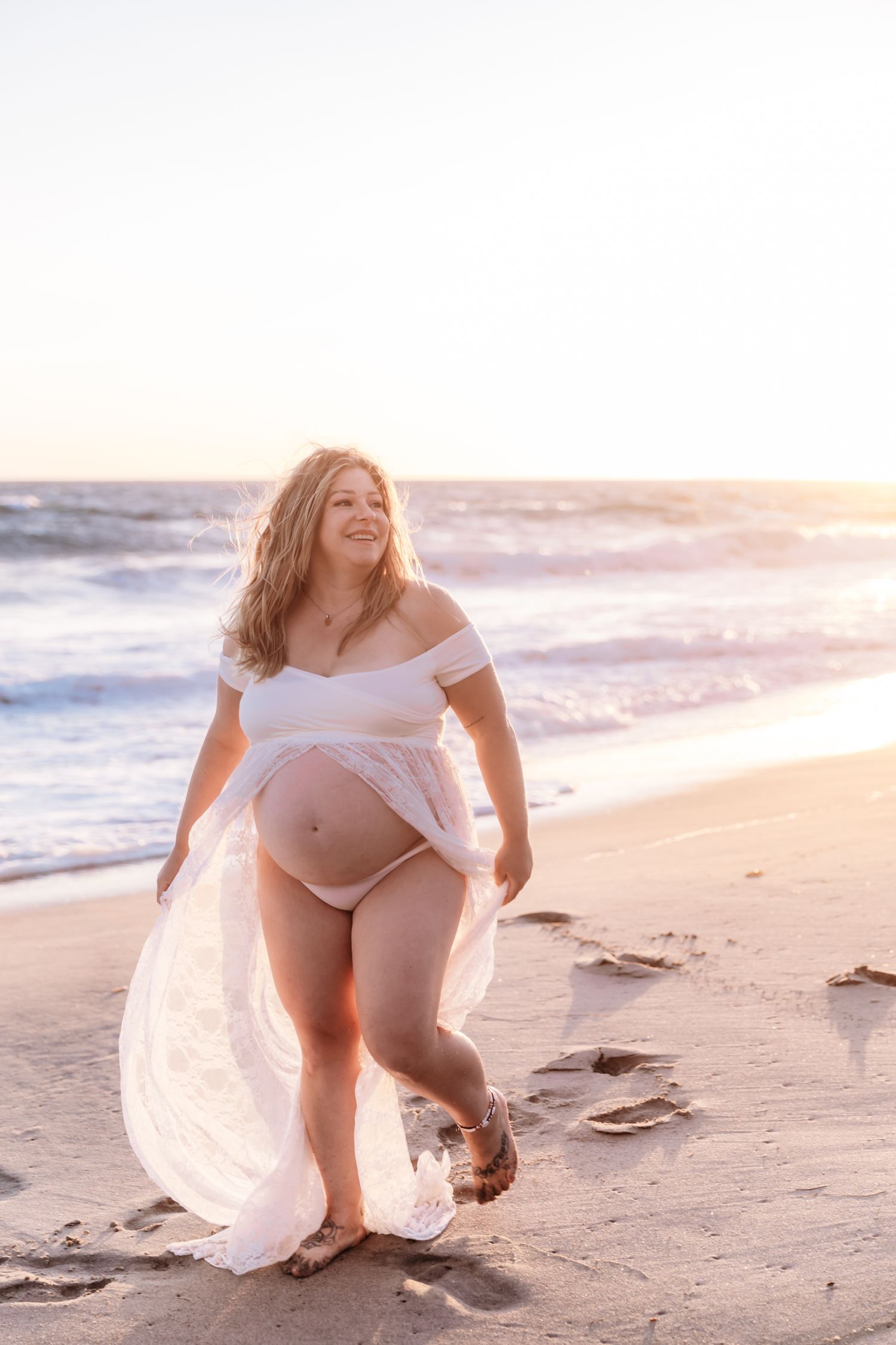 A pregnant woman in a flowing white dress walks along a beach at sunset as waves crash on the shore.