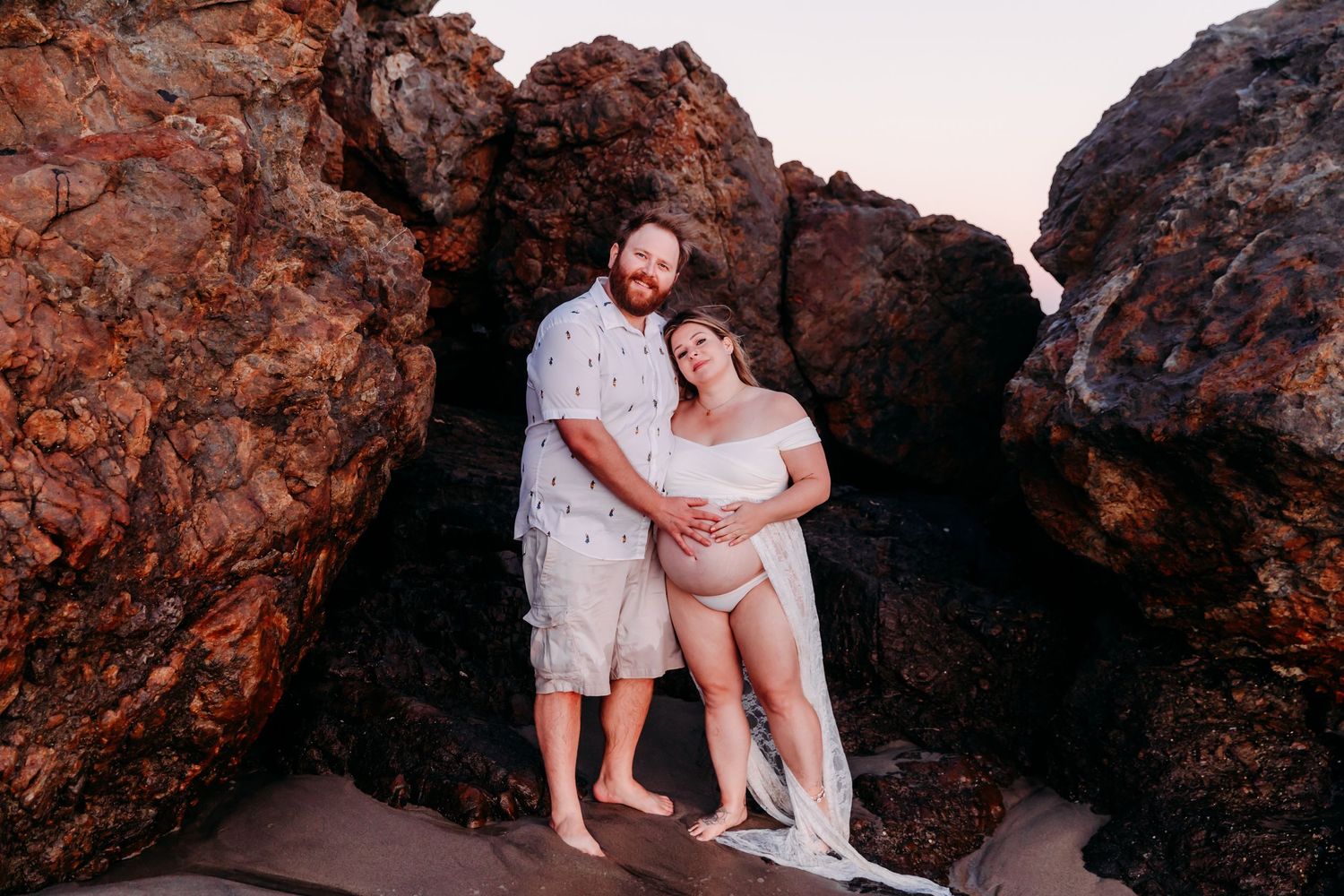 Two people stand together between red rock formations at sunset on a beach.