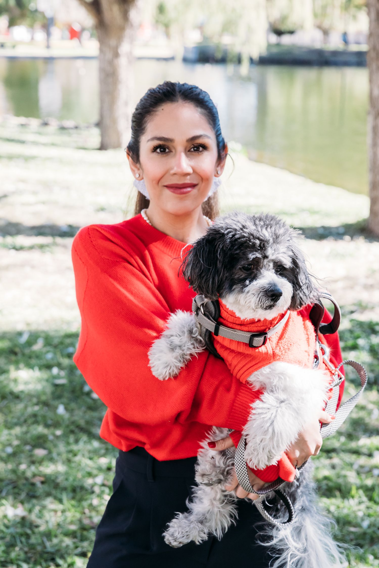 Woman in red sweater holding a fluffy white and grey dog wearing matching red outfit by a lake.
