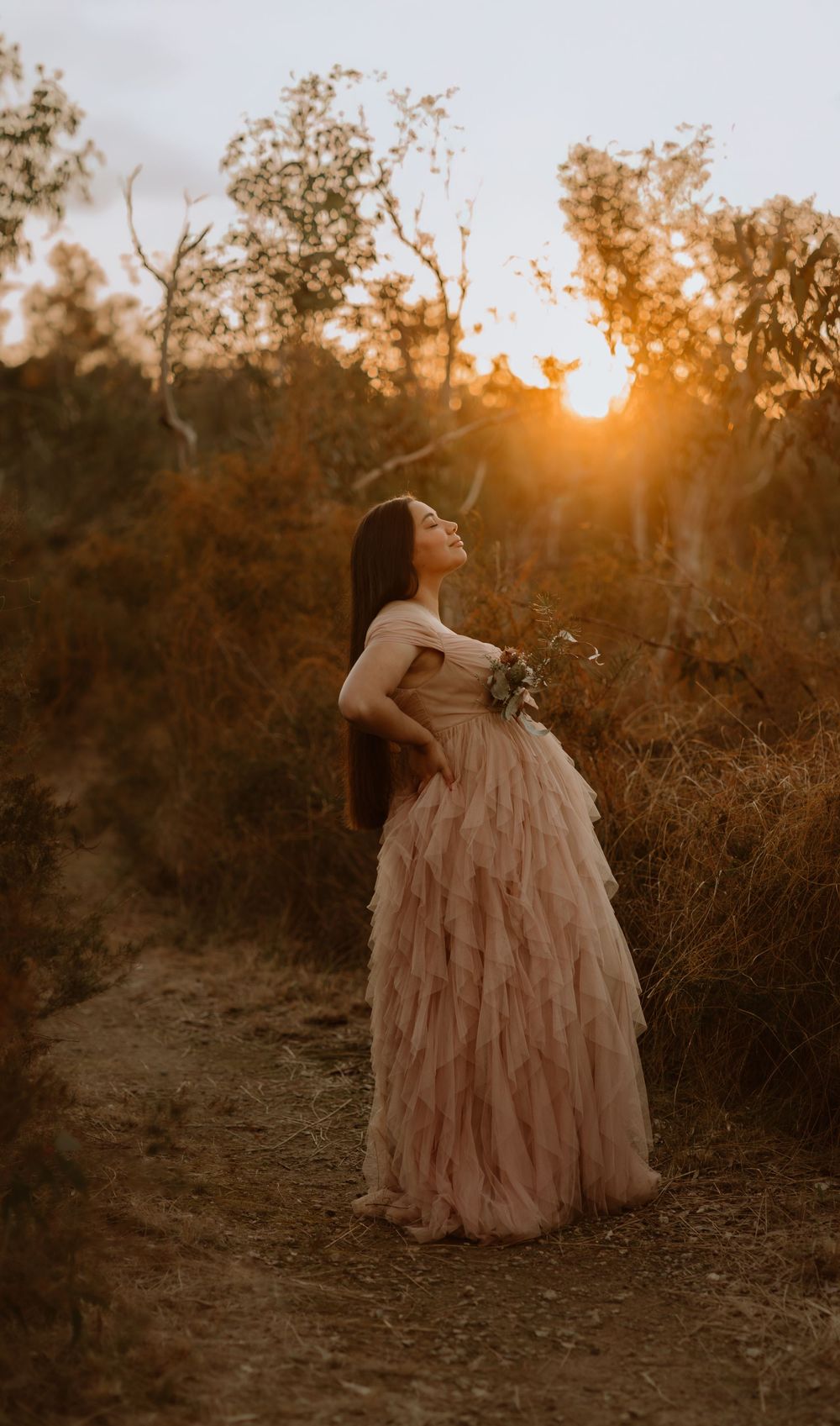 A figure in a flowing pink ruffled gown stands against a golden sunset in a rustic outdoor setting.
