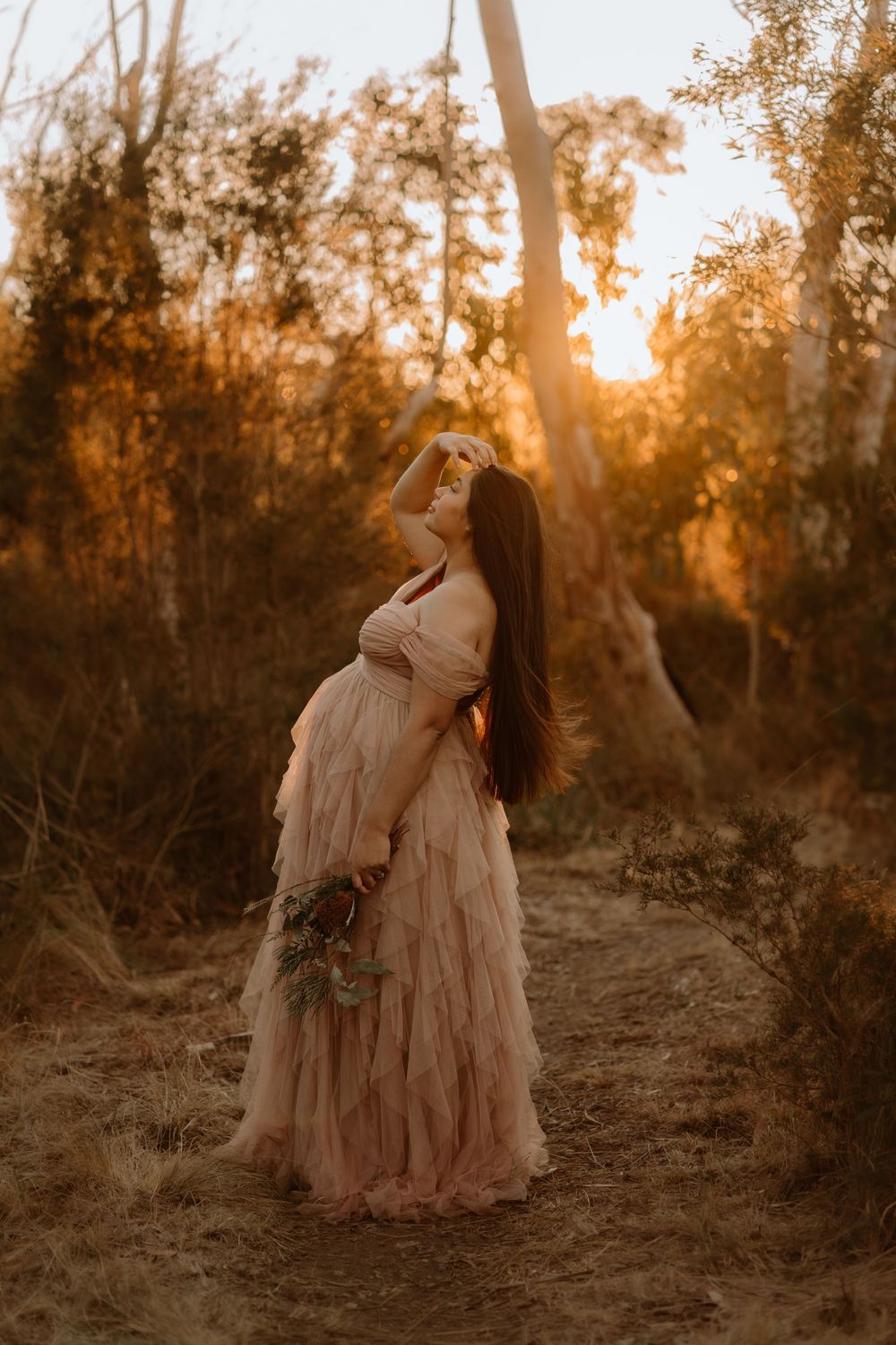 A couple in flowing cream dresses embraces during a romantic sunset photoshoot in a wooded setting.
