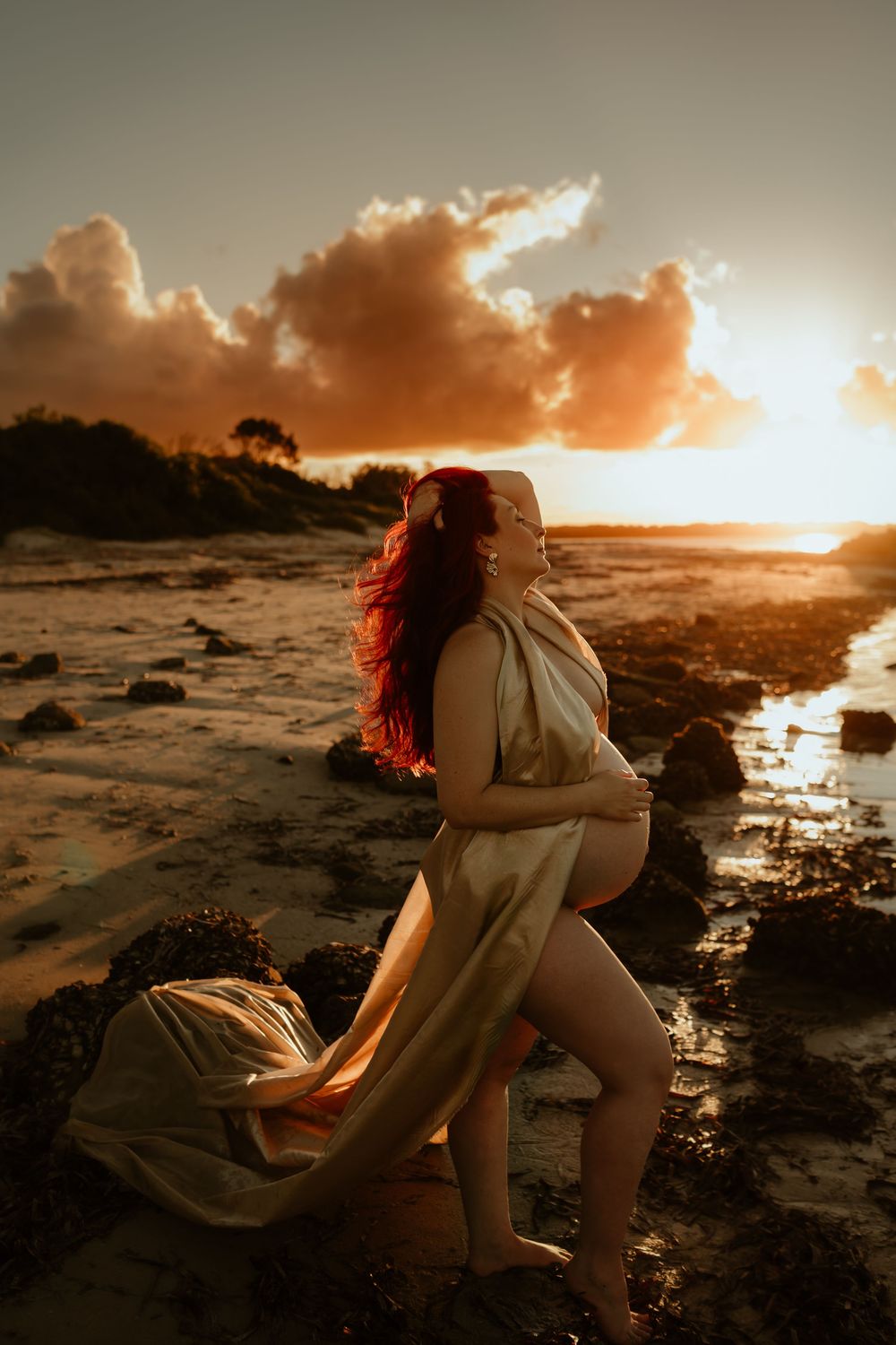 Pregnant woman with red hair draped in flowing fabric poses on rocky beach at golden sunset.