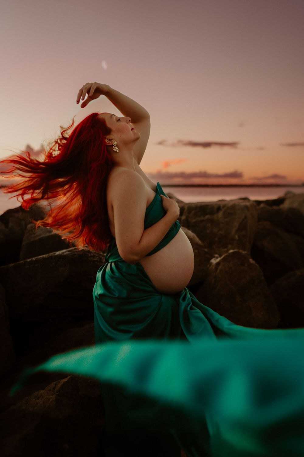 Pregnant woman with flowing red hair poses in a teal gown on rocks at sunset by the ocean.