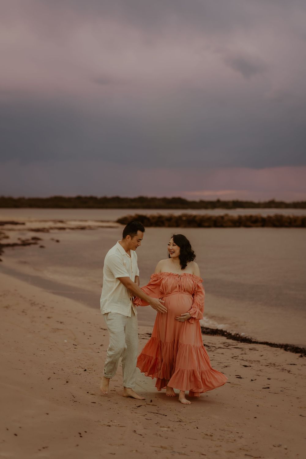 A couple walks hand in hand on a sandy beach at sunset, with the woman wearing a flowing coral dress and man in white.