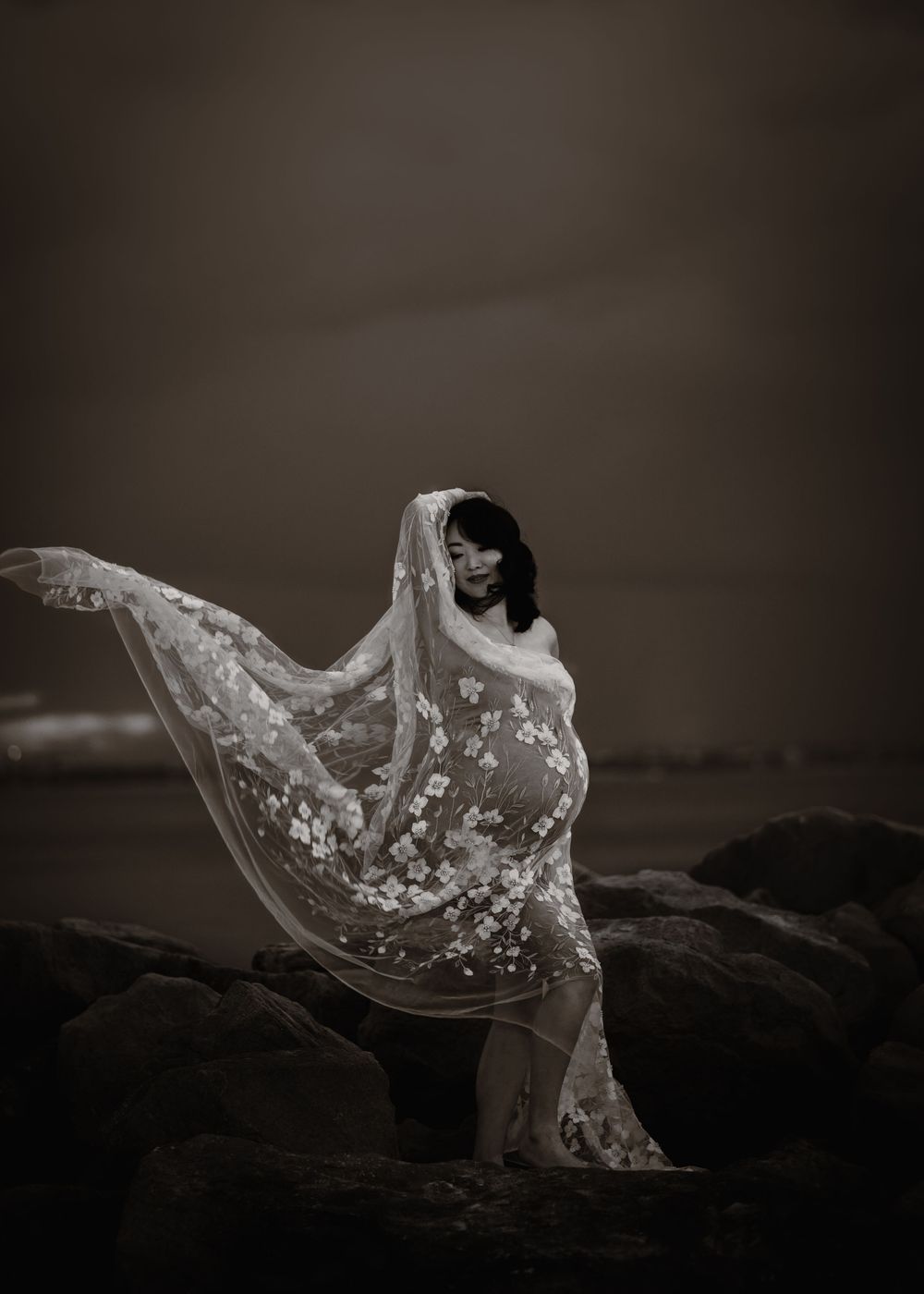 A dramatic black and white photo of a figure on rocks draped in a flowing sheer veil with delicate floral embellishments.