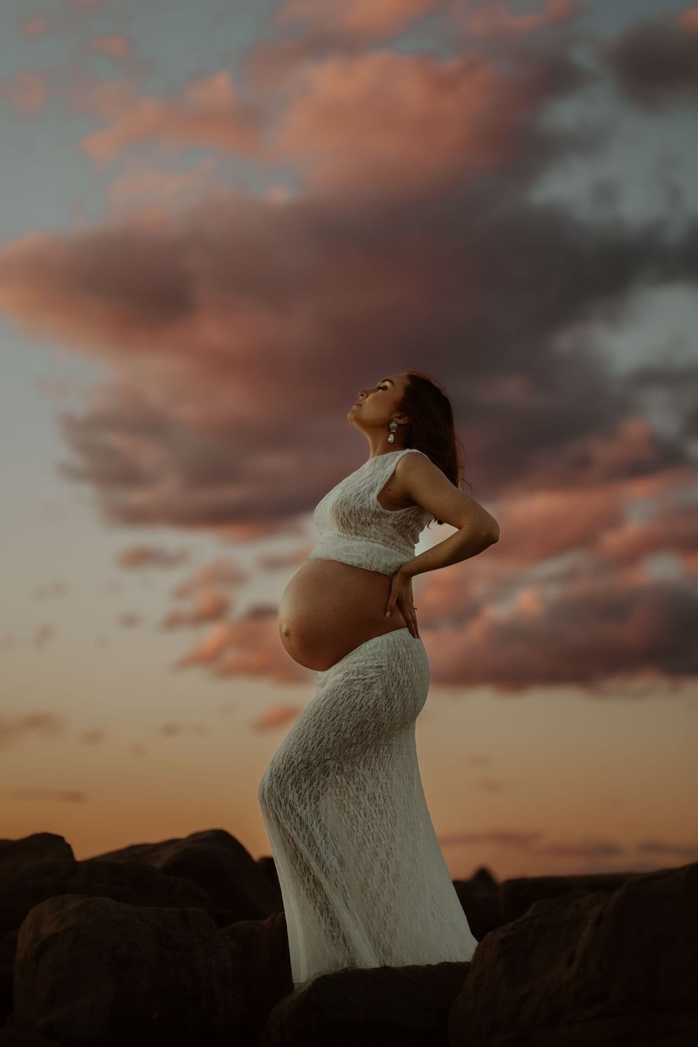 A maternity shoot captures silhouette of a woman in white dress against dramatic orange sunset sky behind rocky landscape.