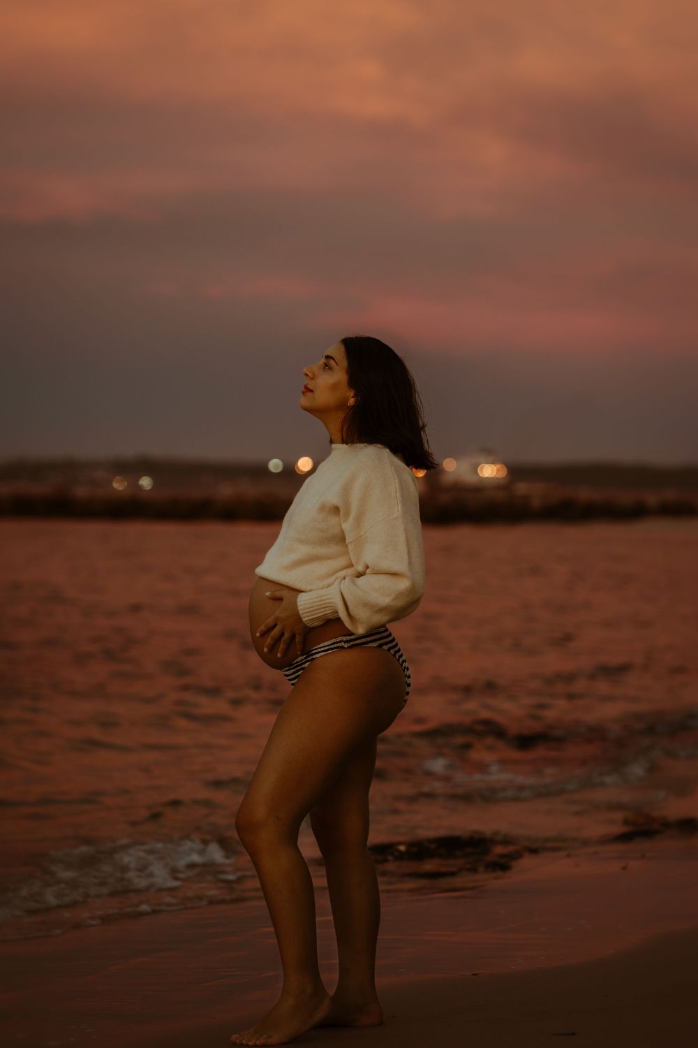 Pregnant woman in white sweater and striped bikini bottoms poses on beach at sunset with pink sky.